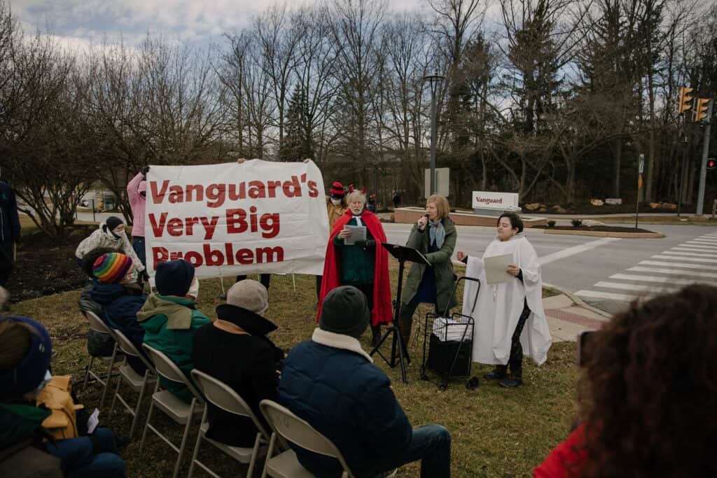 One person speaks into a microphone, while someone dressed as an angel stands to one side of them and someone dressed as a devil stands to the other side. Next to the them of them, three more people hold up a large white banner that says "Vanguard's Very Big Problem"