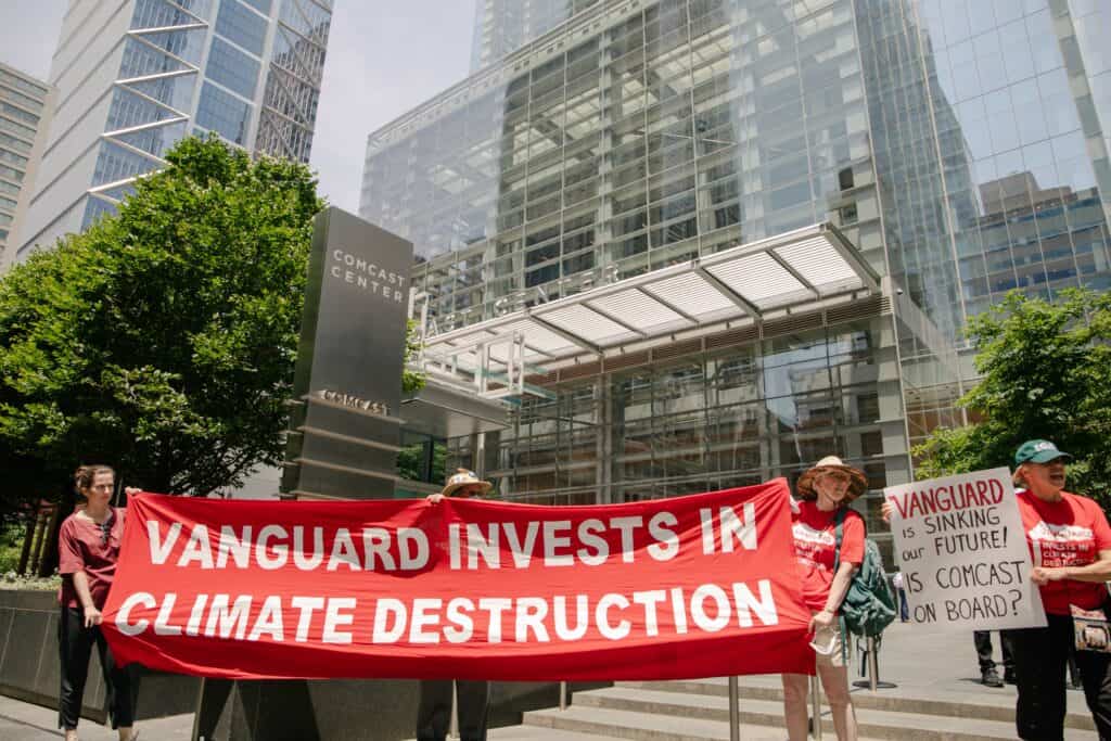 A few people stand in front of a large glass building and a gray sign that says "Comcast Center." Three people are holding a red banner that says "Vanguard invests in climate destruction" and one person is holding a white sign that says "Vanguard is sinking our future! Is Comcast on board?"