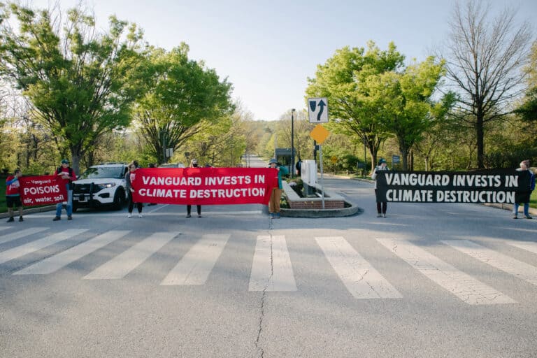 Seven people stand across three lanes of road, holding large red and white banners with white text