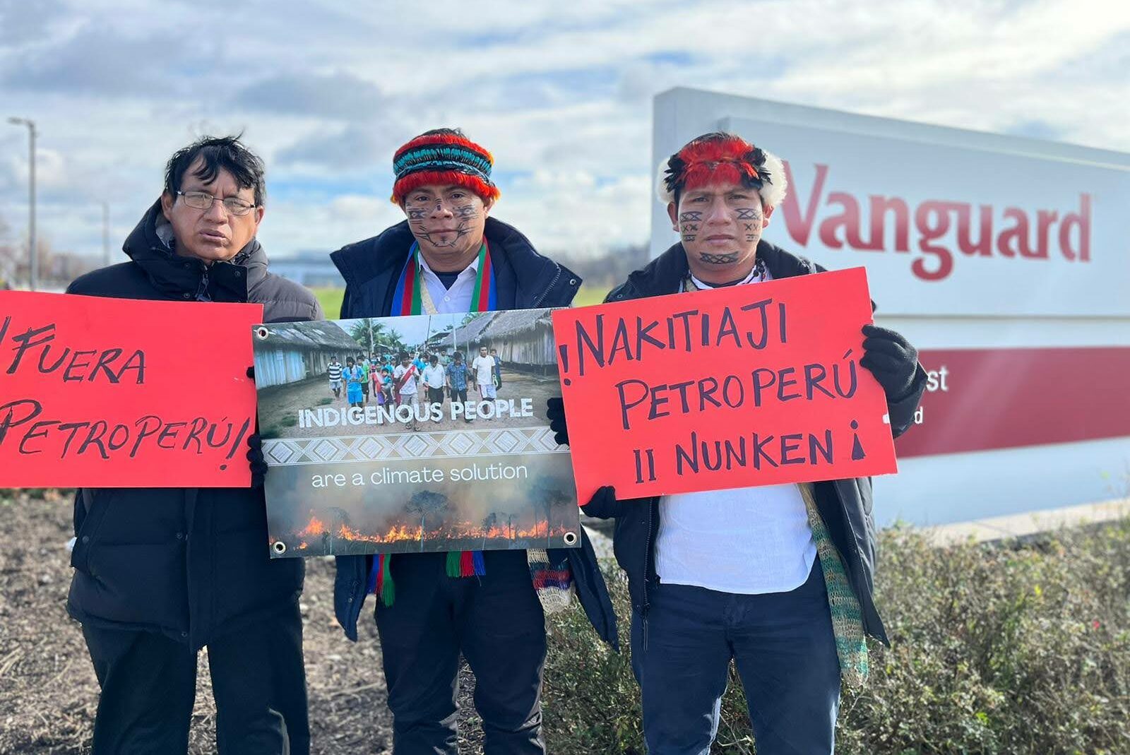 Three people stand at the entrance to Vanguard with signs in English, Spanish, and Wampis. The sign in English says, "indigenous people are a climate solution."