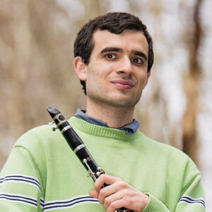 A white person with short, dark hair and a light green sweater holds a clarinet and looks off to the left of the camera