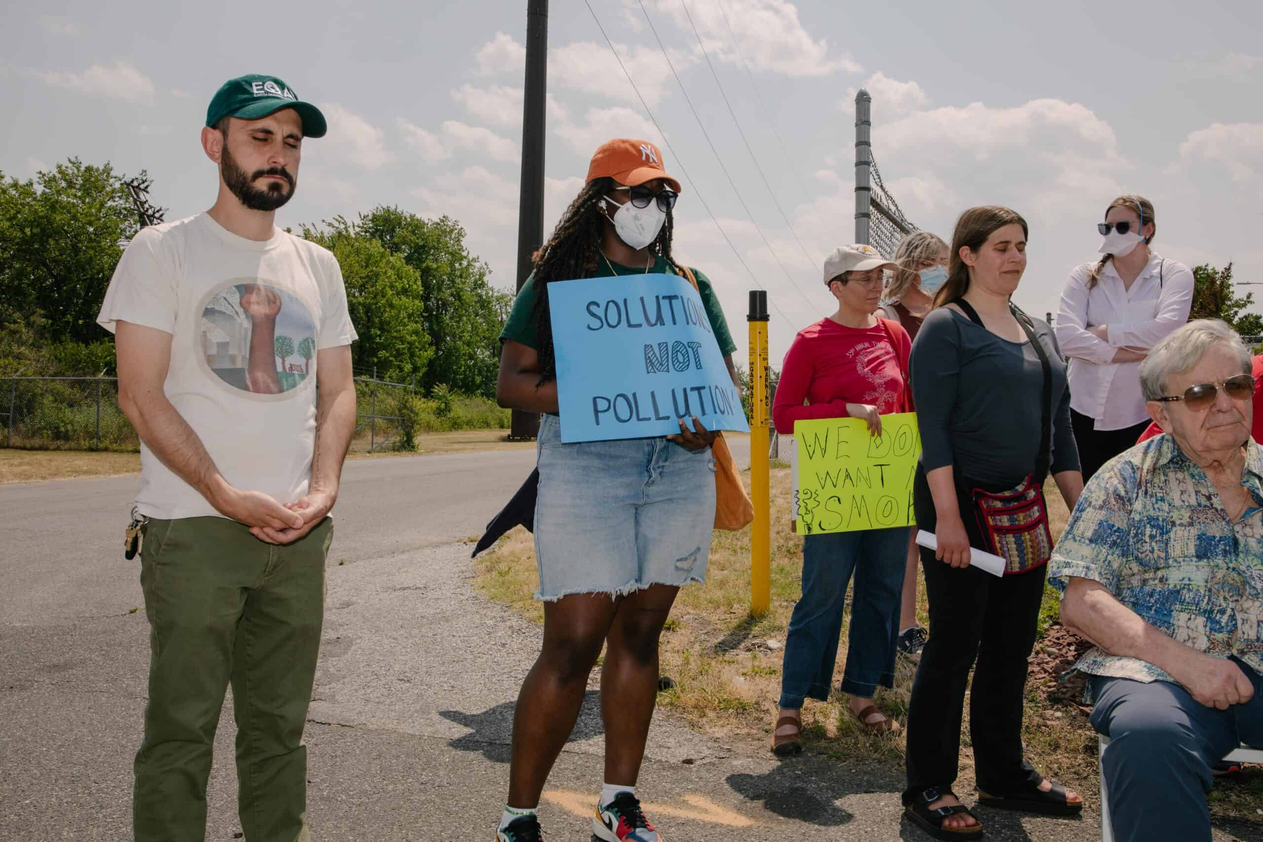 About seven people are seen standing and sitting outside, many with their eyes closed in reflection. One person is holding a sign that says "solutions not pollution."