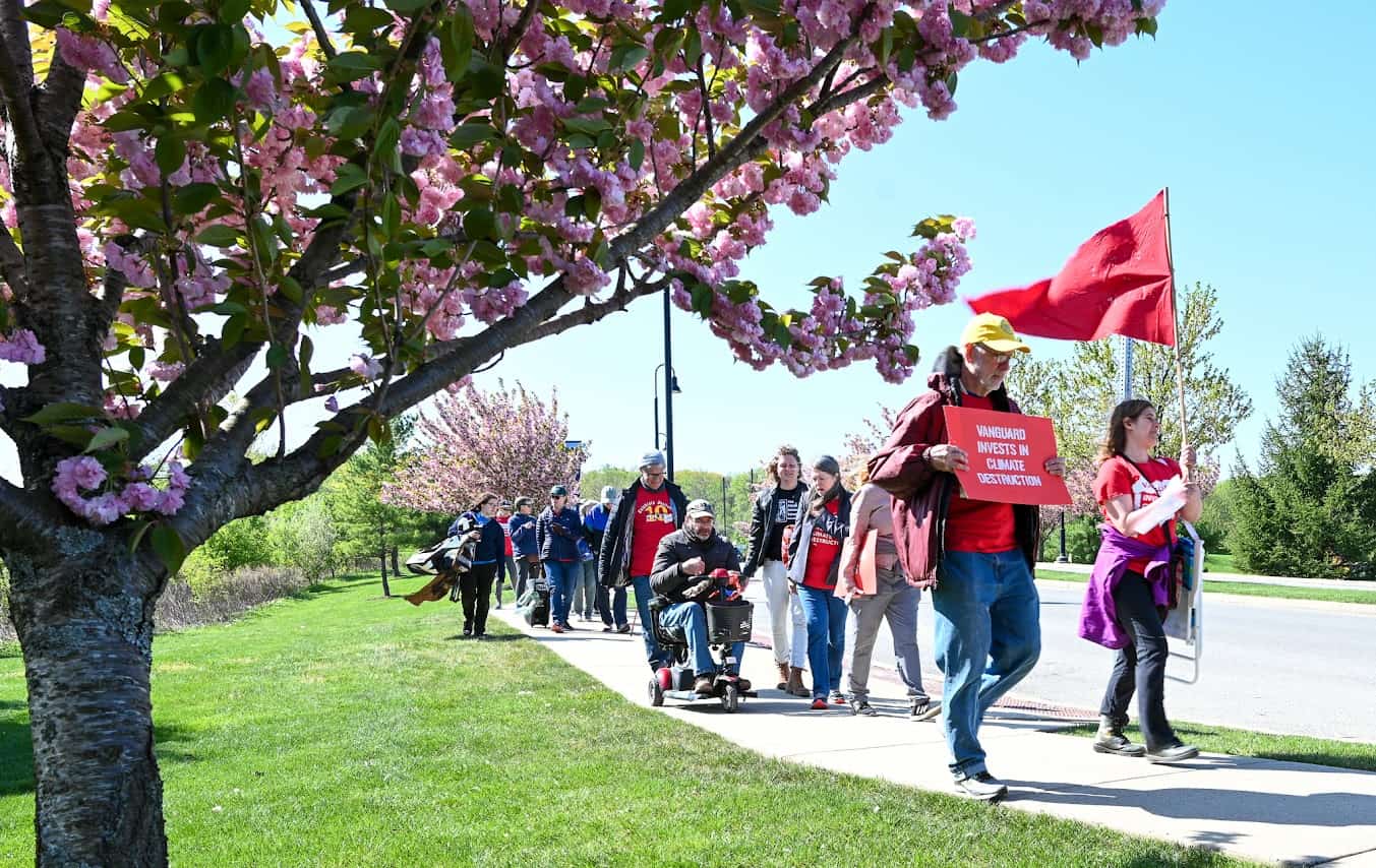 In the foreground, there's a tree with pink flowers and green leaves. In the background, people march down a sidewalk, holding red signs and flags.