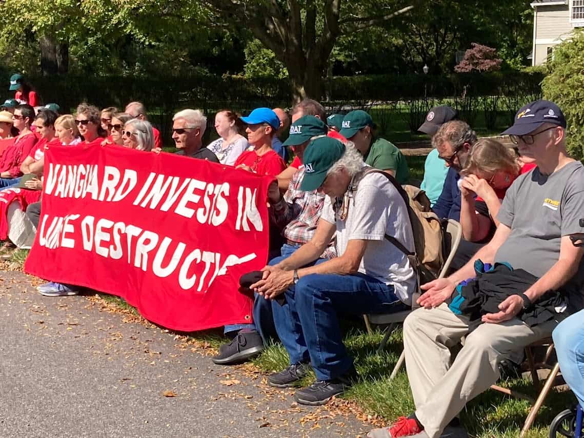 About two dozen people sit in folding chairs outside. Many of them have their eyes closed or are hunched over, contemplatively. Some are holding a large red banner with white text that says "Vanguard invests in climate destruction."