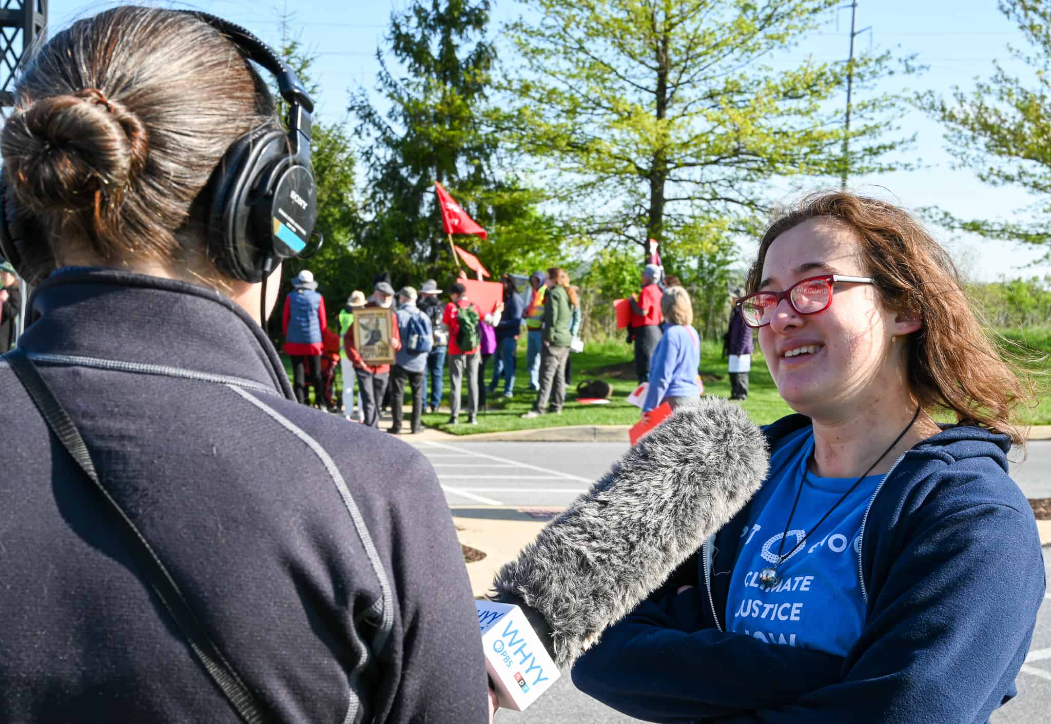 A person wearing glasses and a blue t-shirt speaks into a large microphone held by another person whose back is to the camera. In the distance, there's a crowd of people, some of which are holding signs or flags. 