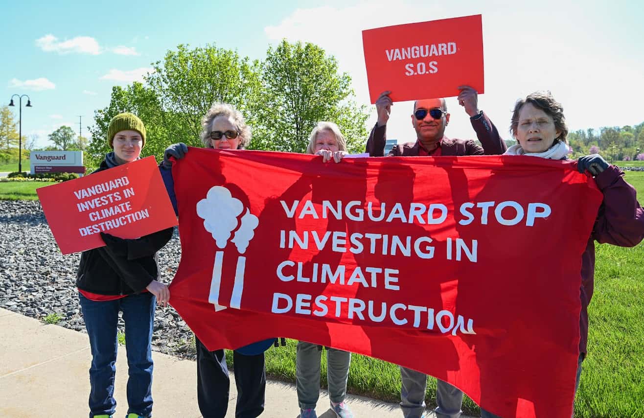 Five people standing outside. Between them, they're holding one red banner with white text that says "Vanguard stop investing in climate destruction" and two red signs with white text that say "Vanguard invests in climate destruction" and "Vanguard S.O.S.," respectively.  