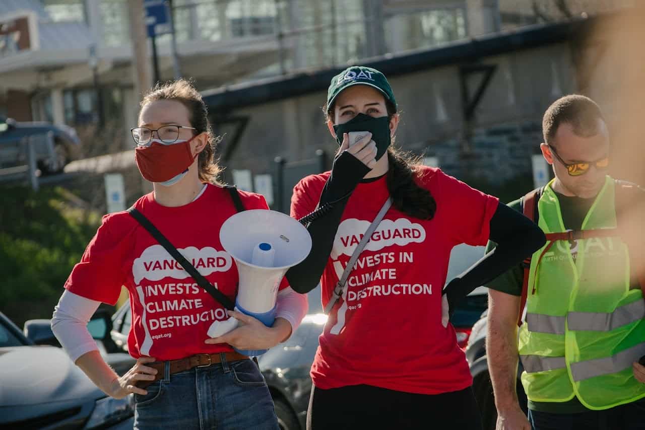 One person in a red shirt with white text that says "Vanguard invests in climate destruction" speaks into a megaphone. A person to one side of them wears a similar shirt and holds the megaphone, while a person on the other side of them wears a neon yellow reflective vest.