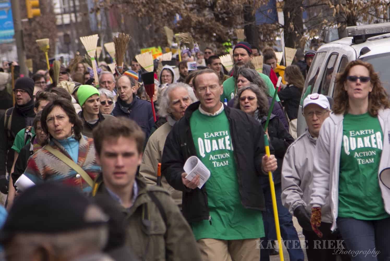 A large crowd of people are marching towards to camera. Many are holding brooms. One person front and center is wearing a green shirt with white text that says "Earth Quaker Action Team."