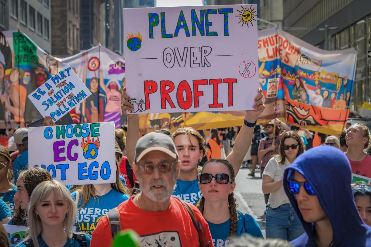Crowd of people in a protest; in the center, there is a person holding up a white sign with colorful text that says "planet over profit"