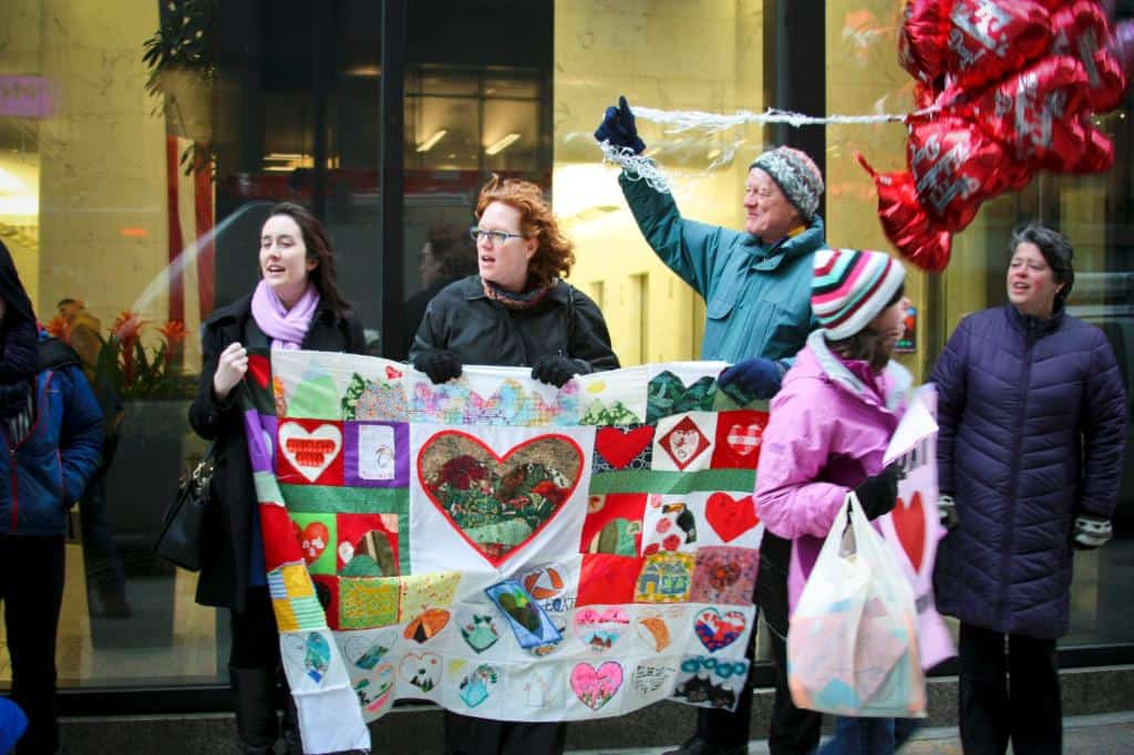 Three people stand outside a glass facade, holding a quilt with various hearts. One of those people also holds a bunch of balloons. 