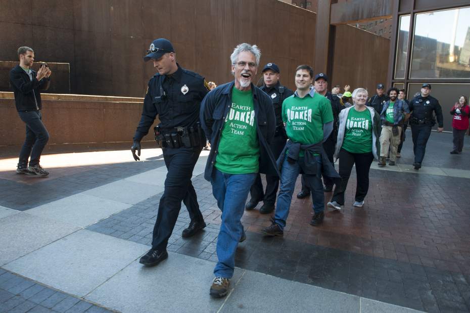 Five people wearing green shirts with white text that says "Earth Quaker Action Team" and with their hands behind their backs are escorted by police officers.
