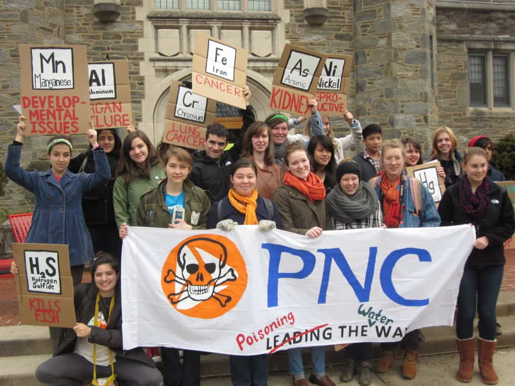 Over a dozen people stand together in front of a stone building to pose for the photo. Many are holding carboard signs over their heads and others are holding a white banner with colorful text and designs. 