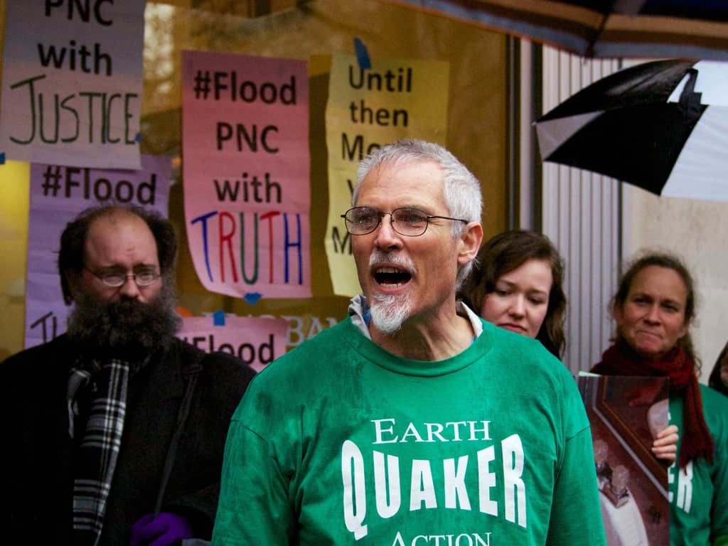 A person in glasses and a green shirt that says "Earth Quaker" is speaking emphatically. Behind them are a few other people and a number of colorful signs that say things like "Flood PNC with Truth" taped to a large window.