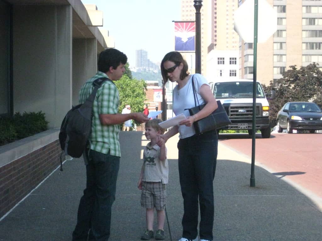 One person hands a flyer to another person holding the hand of a child. They seem to be talking. 