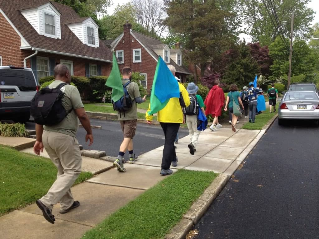 About seven people walk down a sidewalk with their backs to the camera. Two people are holding green and blue flags. 