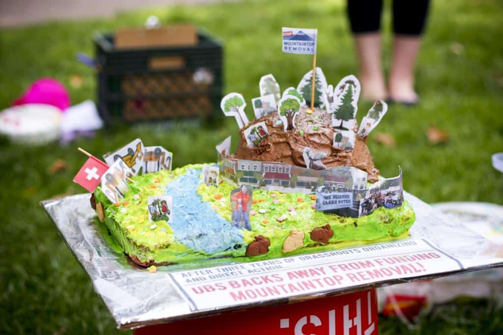 A cake fancifully decorated to look like a wooded mountain, a valley, and a river. On the cake board, there are the words "UBS backs away from mountaintop removal."