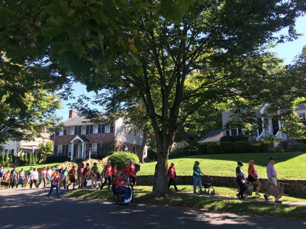 Dozens of people walk down the sidewalk and one rides a motorized scooter, under the shade of a tall tree.