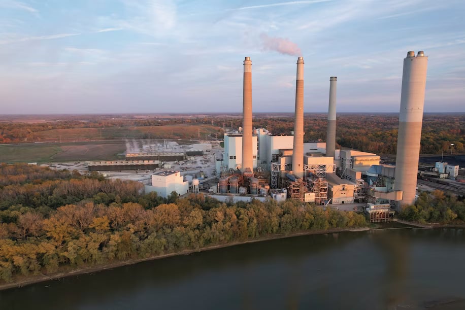 A large industrial complex with four smokestacks along a tree-lined river, with an autumnal landscape stretching off to the horizon