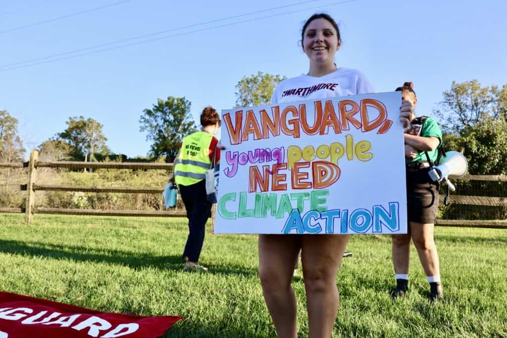 A person stands outside, smiling at the camera and holding a white sign with colorful text that says "Vanguard, young people need climate action." Nearby, there are two other people standing or walking and a red banner with white text lying on the grass.