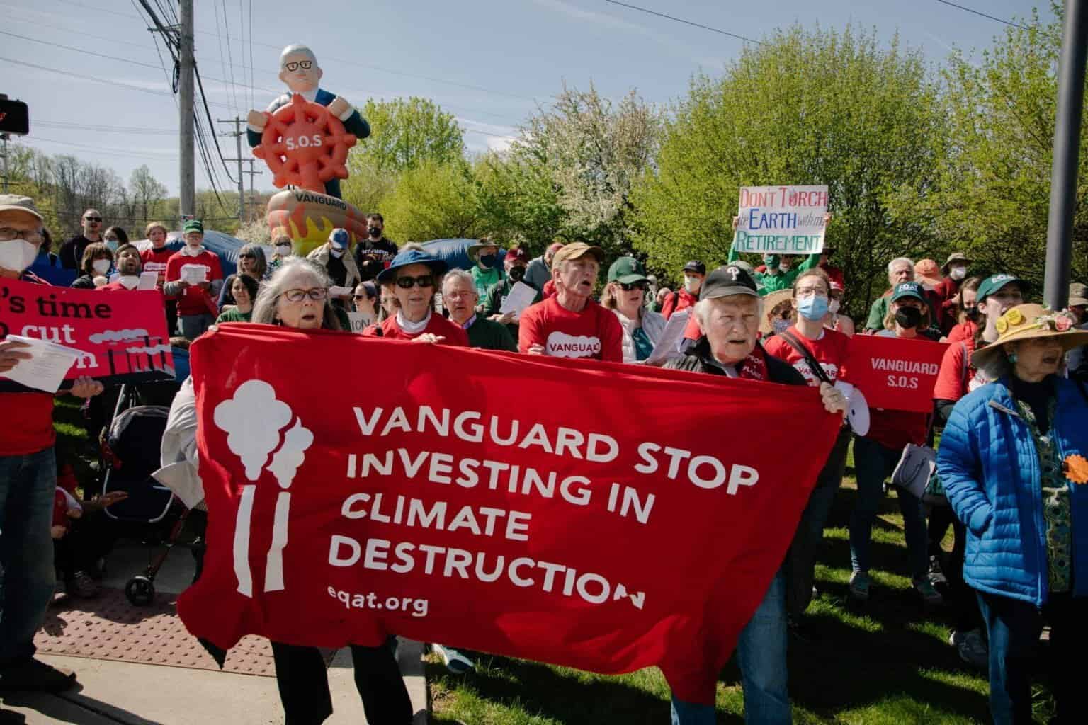 A large crowd of people standing outside, singing. Two people in the foreground are holding a red banner with white text that says "Vanguard stop investing in climate destruction."