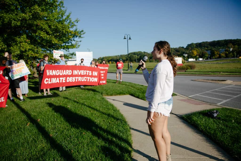 A person in a white long sleeve shirt and jean shorts stands outside, facing left, and speaks into a microphone. Beyond them, there are at least a dozen people holding banners and signs, including a large red banner with white text that says "Vanguard invests in climate destruction."