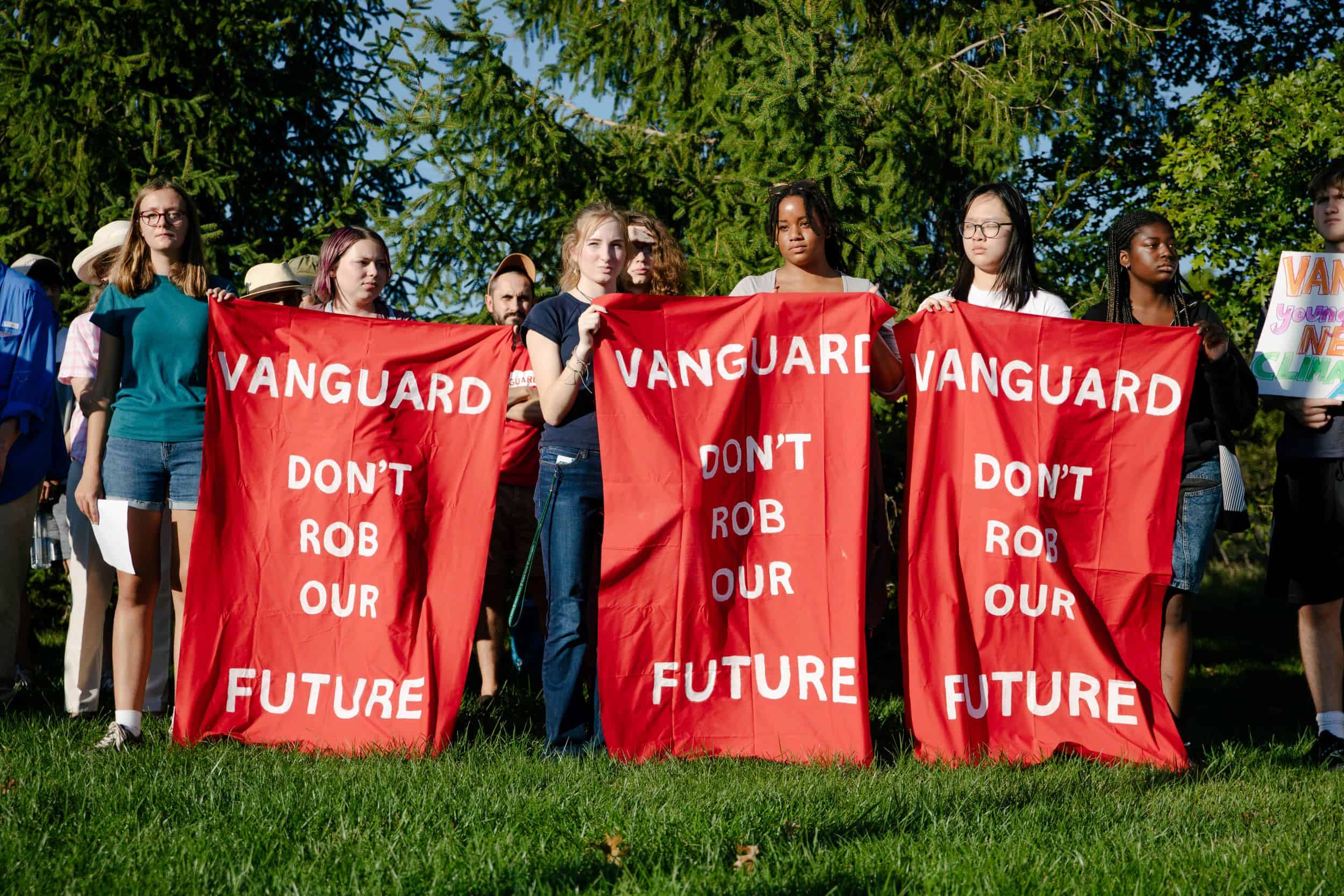 A dozen people stand outside on green grass in front of green trees. 6 people are holding 3 red banners with white text that each say "Vanguard don't rob our future."