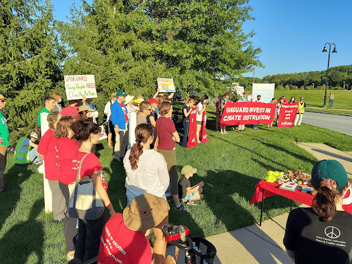 About four dozen people stand outside on green grass, in between green tress and a sidewalk and road. Many people are holding banners and signs. There's a folding table with a red tablecloth and assorted items on top.