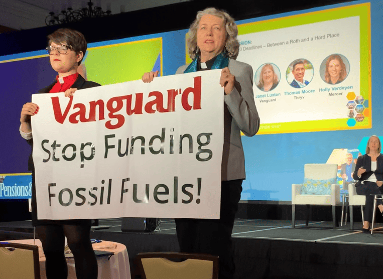 Two people with Roman collars are inside, standing on chairs and holding a banner that says "Vanguard Stop Funding Fossil Fuels!" Behind them is a person sitting in a chair on a stage and a large screen showing photos and names of presenters.