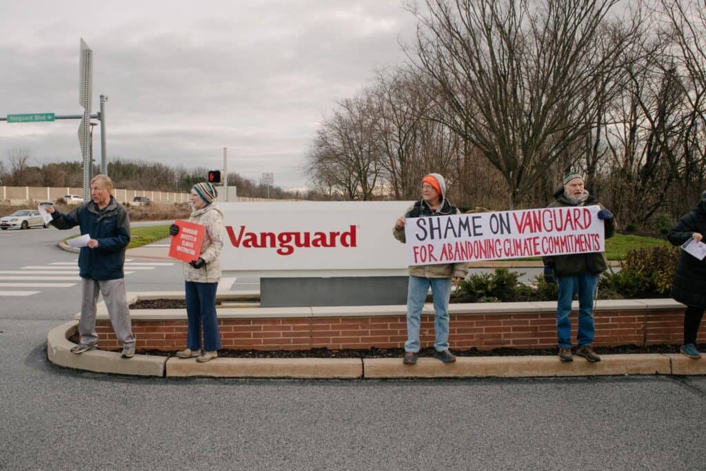 Five people stand on the curb, in between a sign that says Vanguard and the road. Two people are holding a banner that says "Shame on Vanguard for abandoning climate commitments."