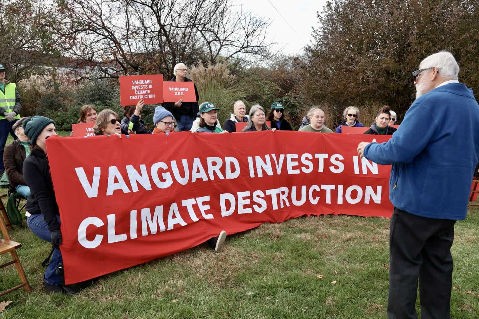 About two dozen people sit on chairs in two rows outside. Those in the first row hold a large red banner with white text that says "Vanguard invests in climate destruction."