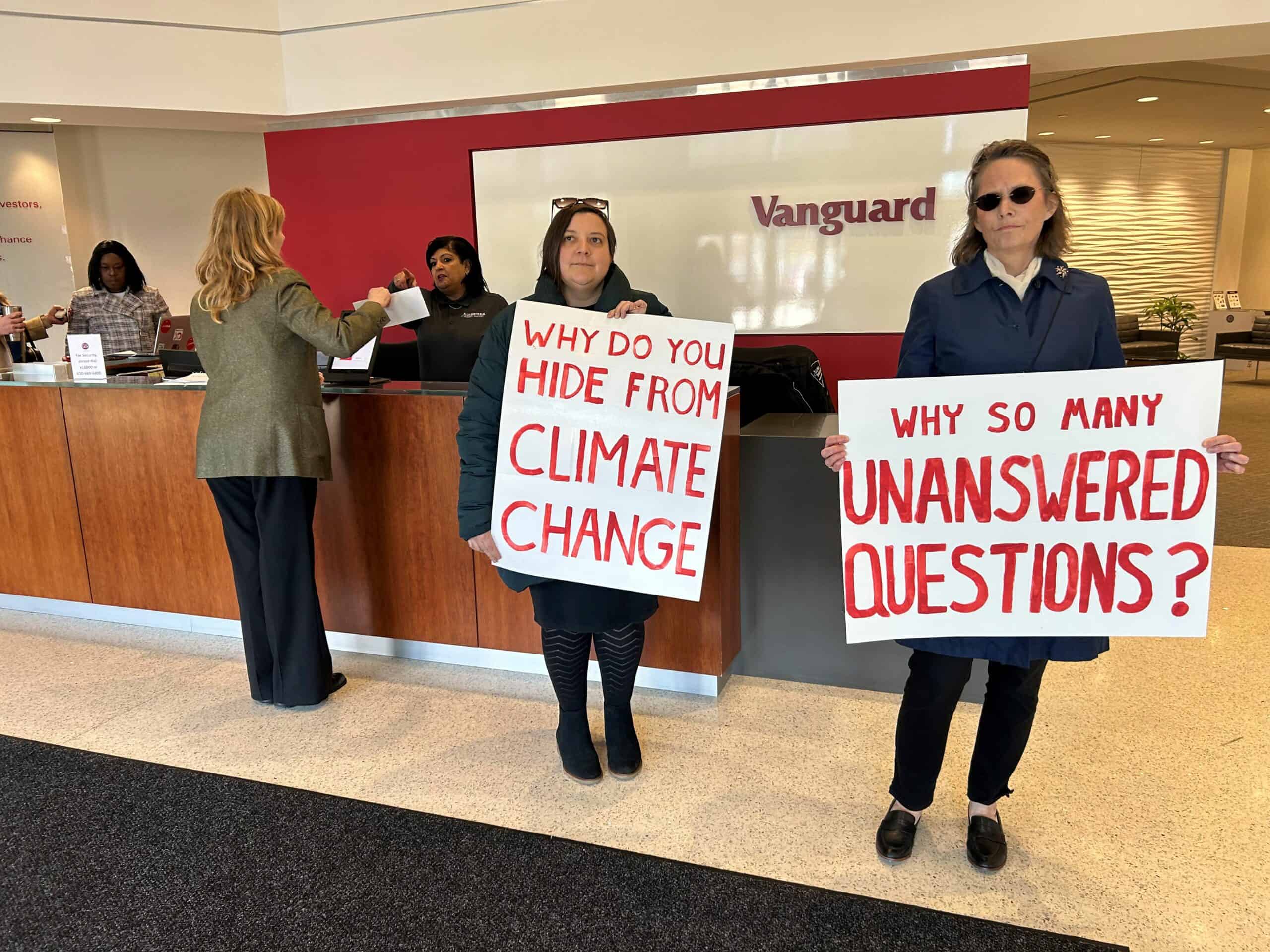 Inside a Vanguard office lobby, two people hold white signs with red text while another person hands a letter to a receptionist who is pointing.