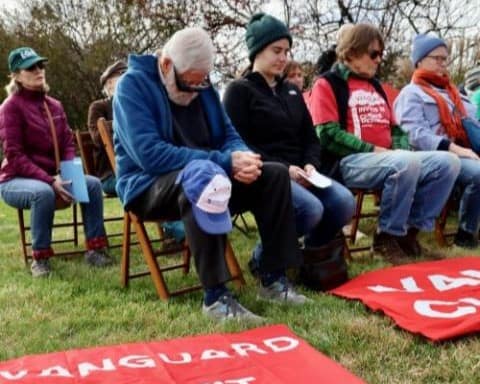 People sit in two rows of wooden folding chairs outside, with their eyes closed or heads bowed. Laying in front of them in the grass are red fabric banners with white text.