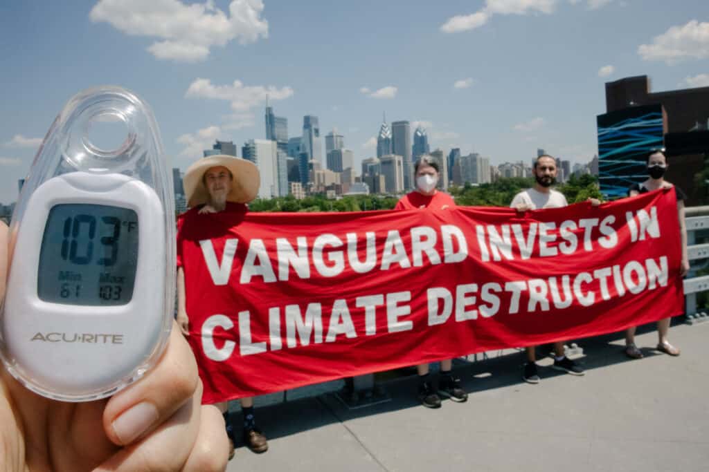 In the foreground, a hand holds up a thermometer that reads 103 degrees, while four people stand behind a banner that says "Vanguard invests in climate destruction"