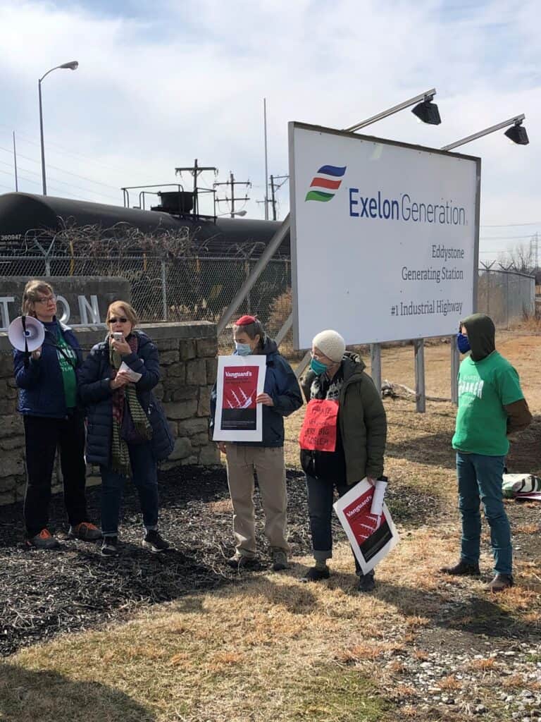 A few people stand outside, in front of a barbed wire fence, a train car, and a sign that says "Exelon Generation Eddystone Generating Station." Two of them are holding signs and one is speaking into a megaphone's microphone.