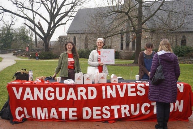 Outside, there's a table with a large red banner draped across it. The banner says "Vanguard invests in climate destruction." Three people are standing behind the table and one person is standing in front of it, facing the others. 