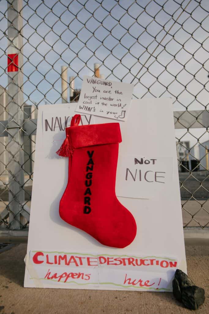 A sign that says "Naughty, not nice / Vanguard / Climate destruction happens here" leans against a chain link fence. There are smokestacks in the background.