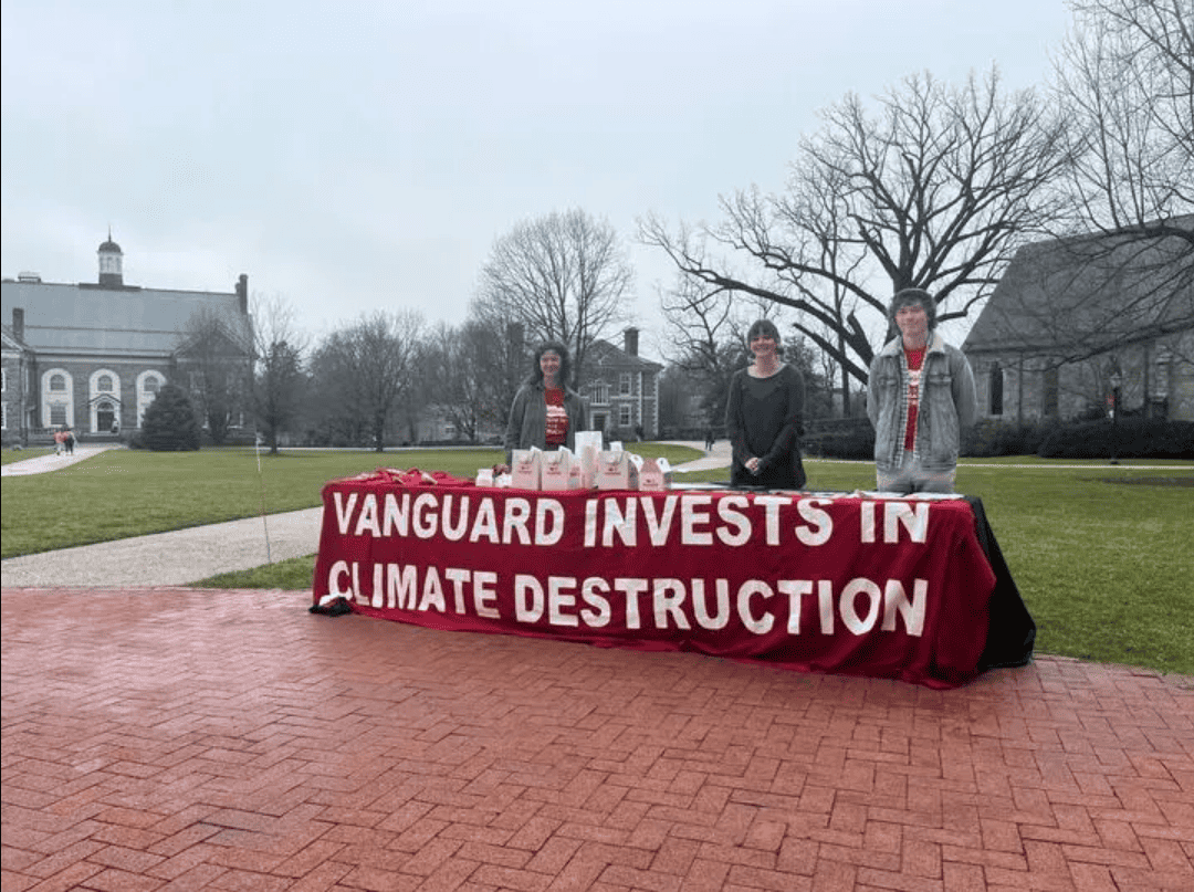 Outside on a cloudy day, three people stand behind a table, draped with a large red banner with white text that says "Vanguard invests in climate destruction"