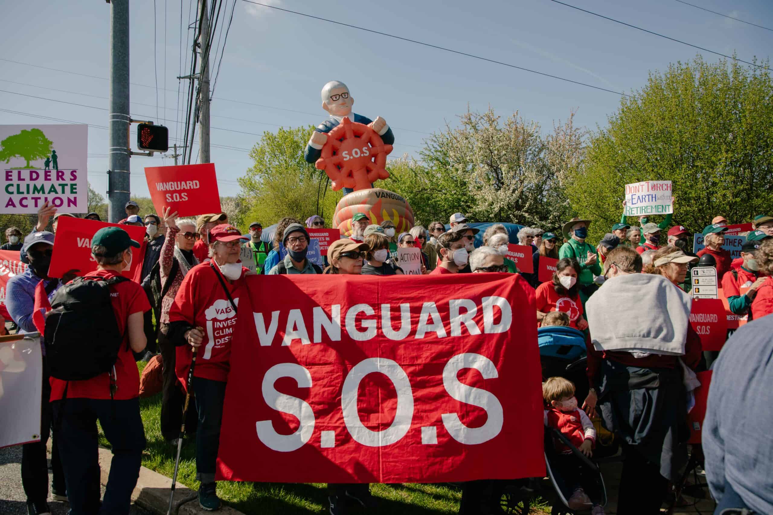 A large crowd of people outside with green trees in the background with a red banner being held saying "Vanguard S.O.S"